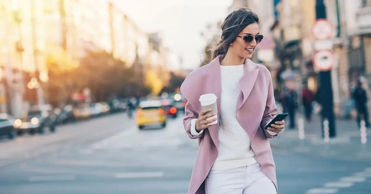 Woman walking with coffee, looking at phone, illustrating a modern professional for 1094 tax form.