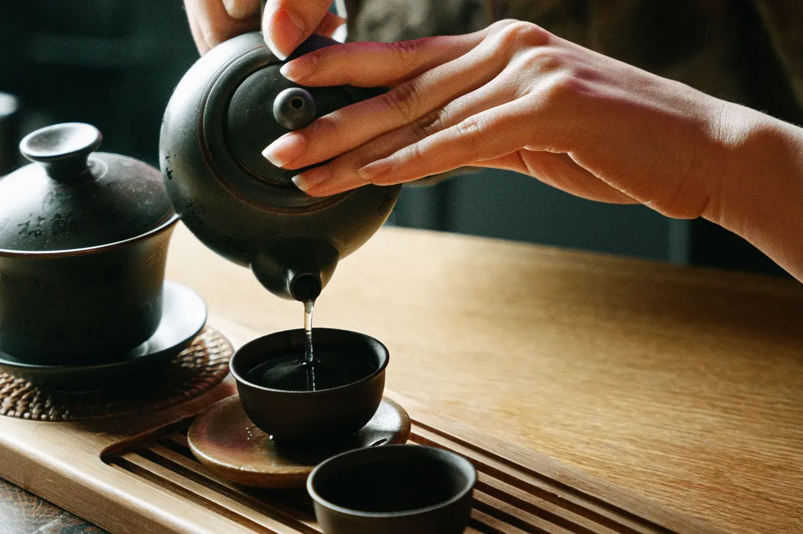 Hands pouring tea from a black clay teapot into a small teacup.