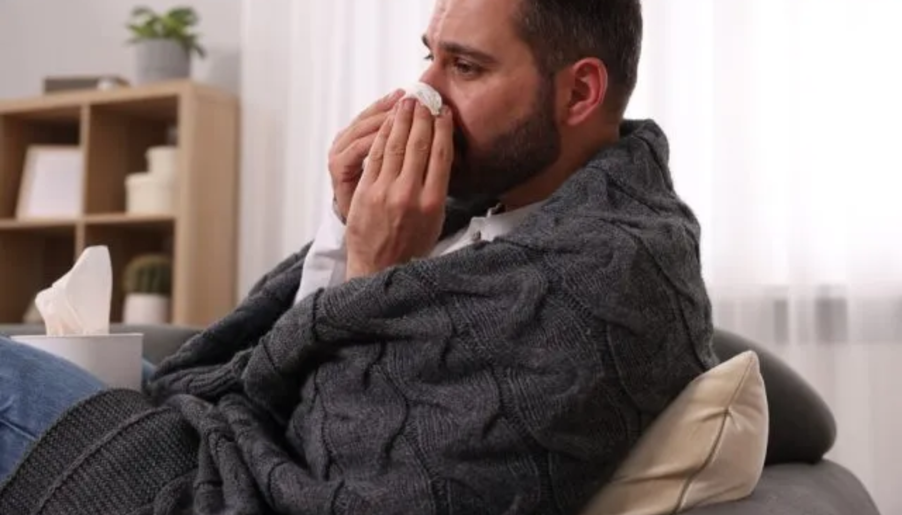 En mann med skjegg sitter på sofaen, innpakket i et teppe, og tørker nesen med et papir mens han ser syk ut.
