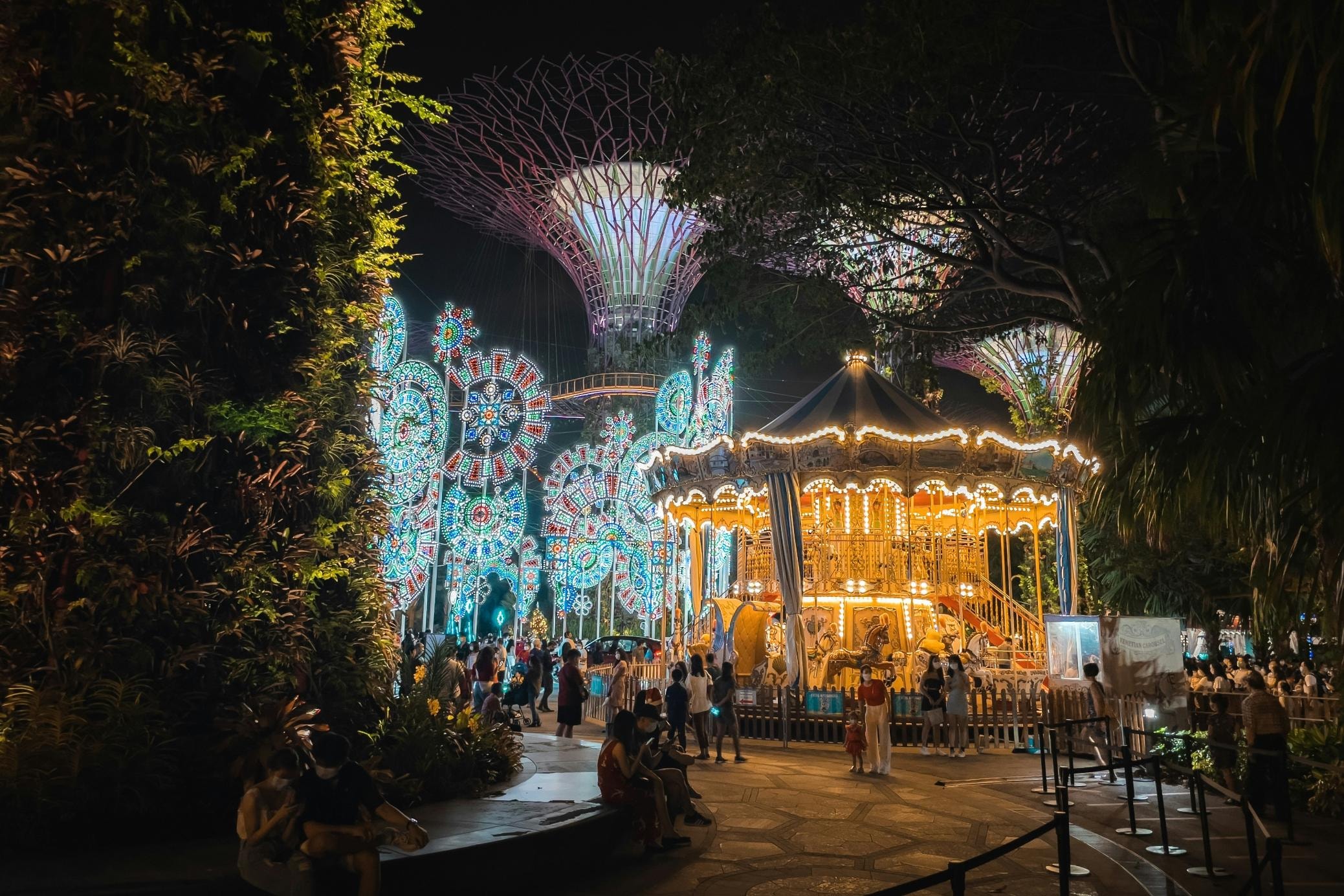 A glowing, classic carousel serves as a focal point in this festive night scene, set against the backdrop of the iconic, illuminated Supertrees at Gardens by the Bay. Visitors stroll through the park, surrounded by intricate light sculptures and lush greenery that enhance the dazzling and magical atmosphere.