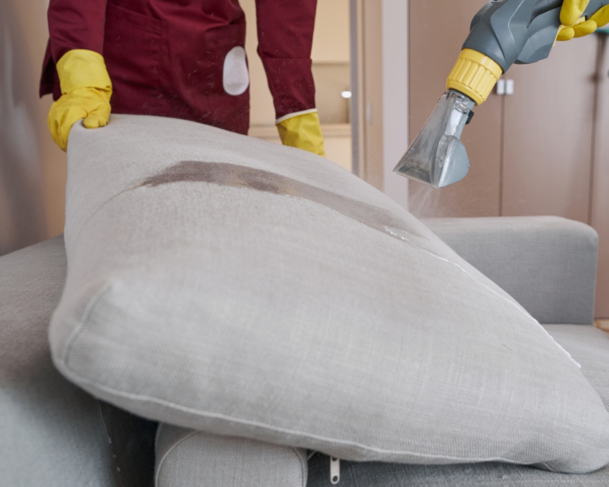 Professional upholstery cleaner using a handheld extraction tool on a light gray sofa cushion, with visible dirt being removed and the fabric being deep cleaned.