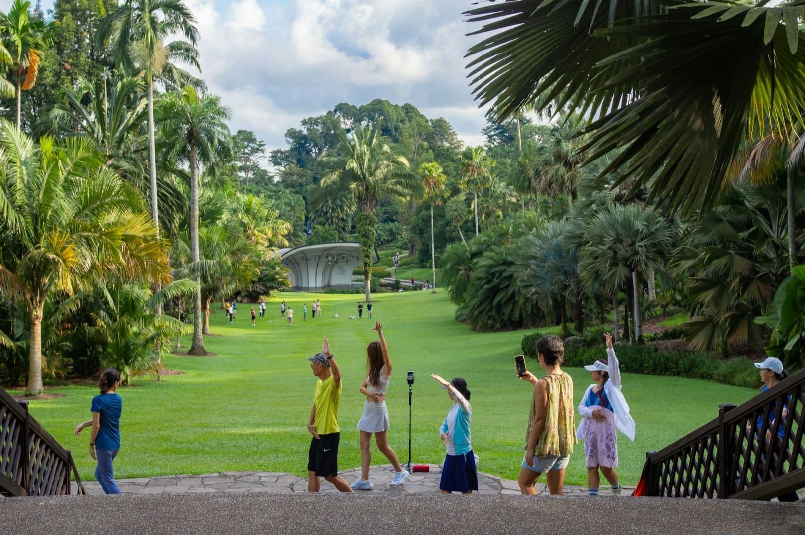 A group of people walks through a lush park with tall palm trees and a distant amphitheater. They appear joyful and relaxed, some holding phones.