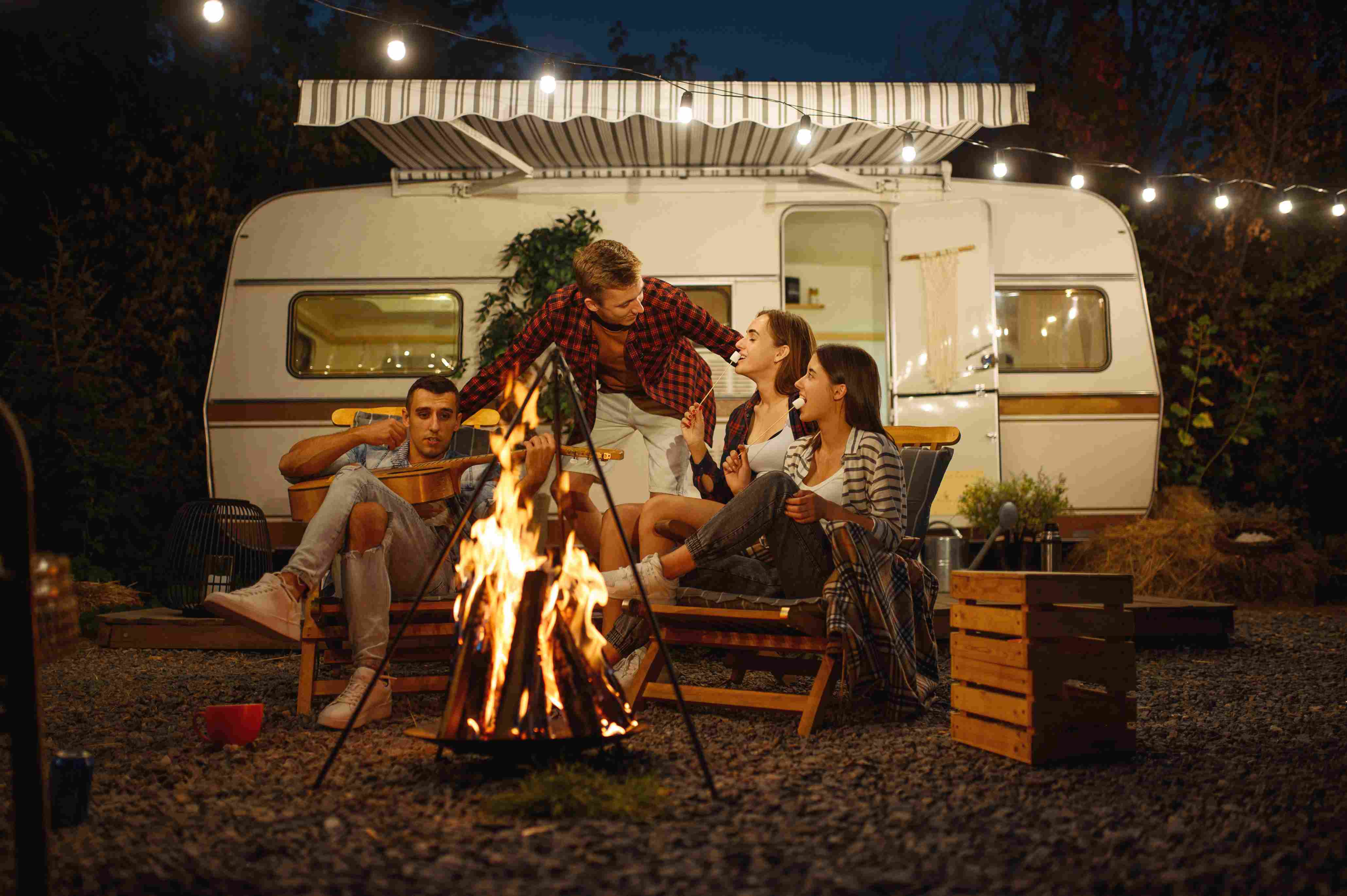 Vier Freunde sitzen am Lagerfeuer vor einem Camper und essen Marshmallows