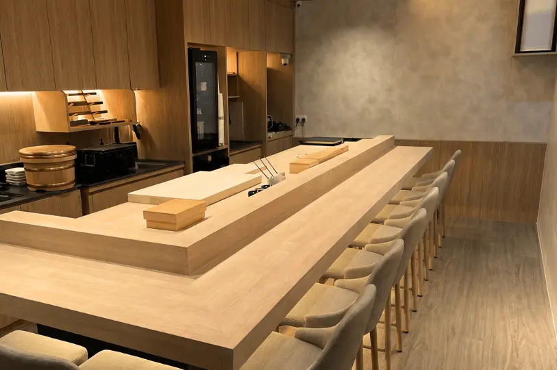 A minimalist, modern Omakase sushi bar counter made of light-colored wood, beautifully arranged with high chairs, clean workstations, and soft ambient lighting.