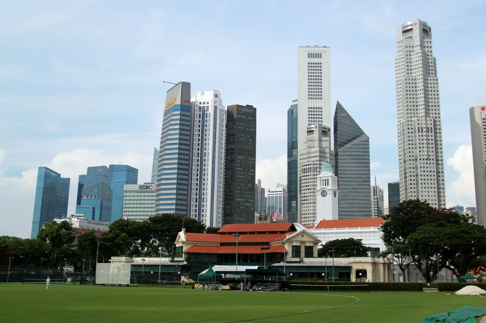A lush green field is in the foreground with a historic building behind it, set against a striking backdrop of modern, high-rise skyscrapers under a clear sky.