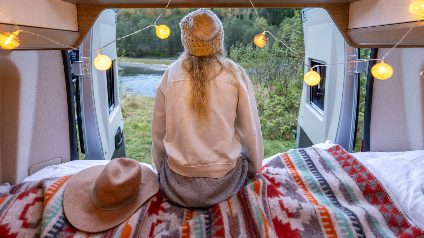Frau sitzt im Camper auf einer bunten Decke und blickt durch die offene Tür auf einen Fluss inmitten grüner Natur, umgeben von Lichterkette