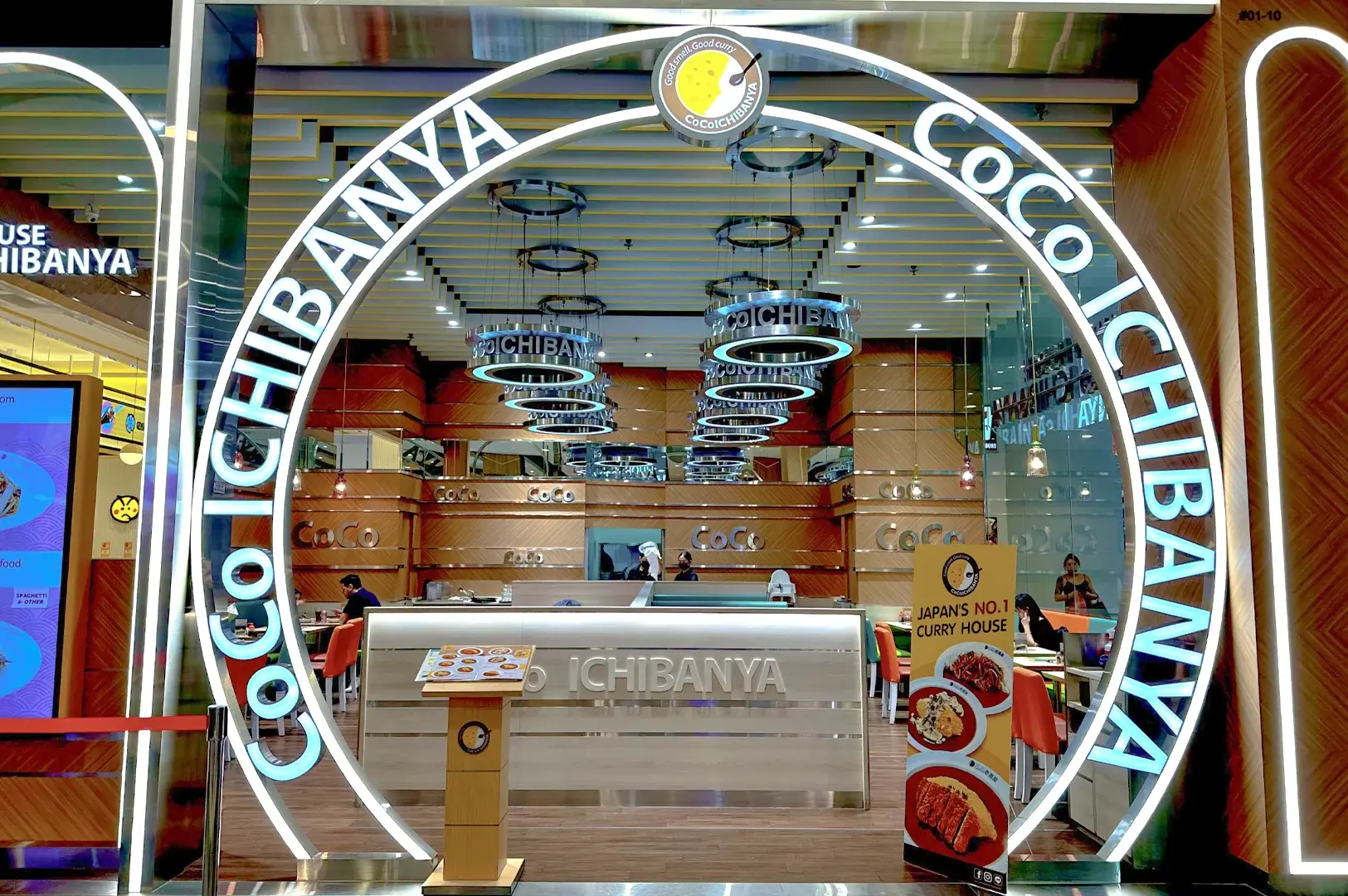 Front-facing eye-level wide shot of CoCo Ichibanya Japanese curry restaurant entrance featuring a glowing circular arch sign, branded “COCO ICHIBANYA” lettering, modern wood panel interiors, and visible dining area for SEO-friendly “CoCo Ichibanya restaurant entrance modern Japanese curry house interior design.