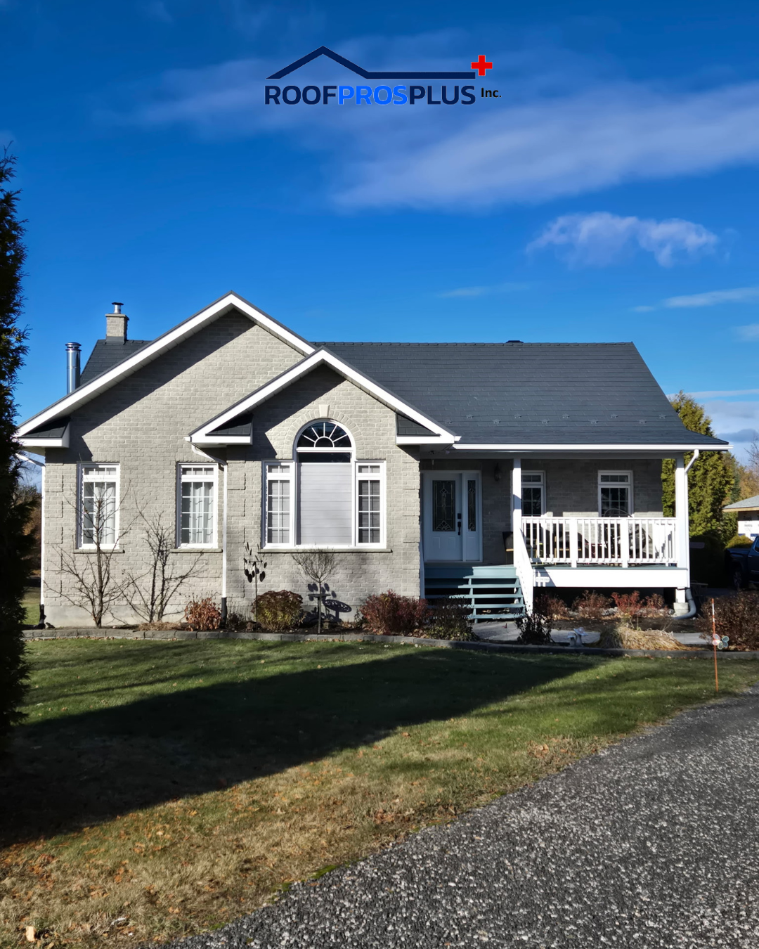 Single-story gray brick house with a dark roof and white trim. A porch with a railing is visible. The foreground shows a grassy lawn under a blue sky with the Roof Pros Plus logo above the home.