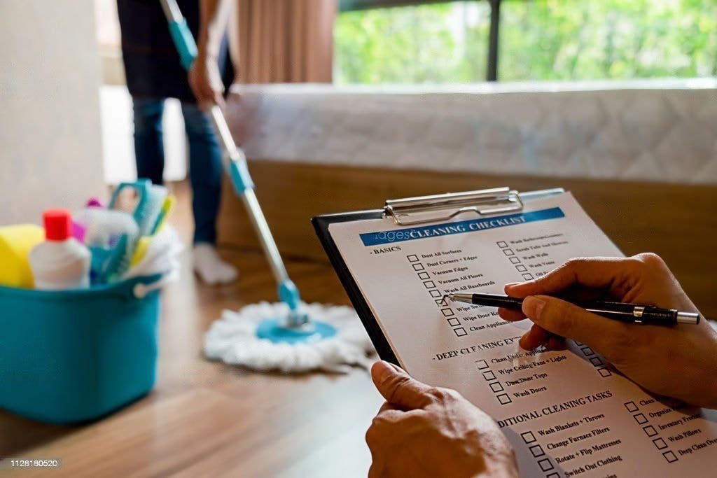 A close-up of a person holding a clipboard and checking items off a cleaning checklist, while another person mops a wooden floor in the background. A blue bucket of cleaning supplies sits nearby.