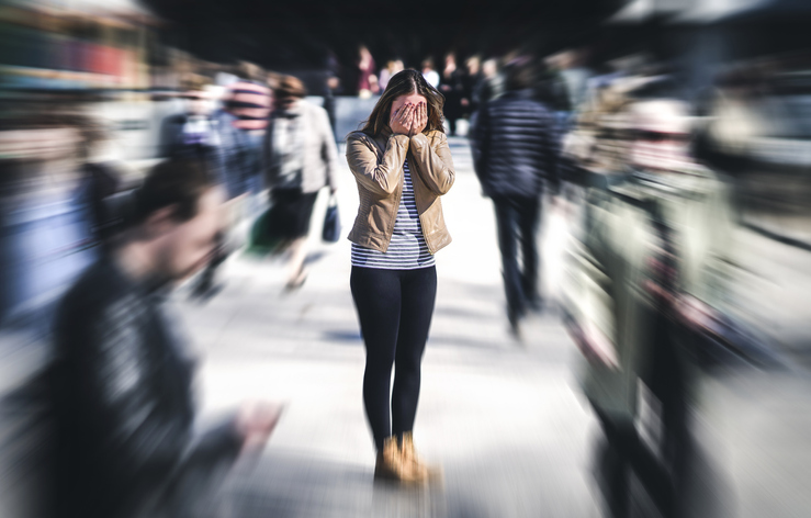 An image of an anxious woman on a crowded sidewalk covering her face with her hands as blurred images of people pass by. 