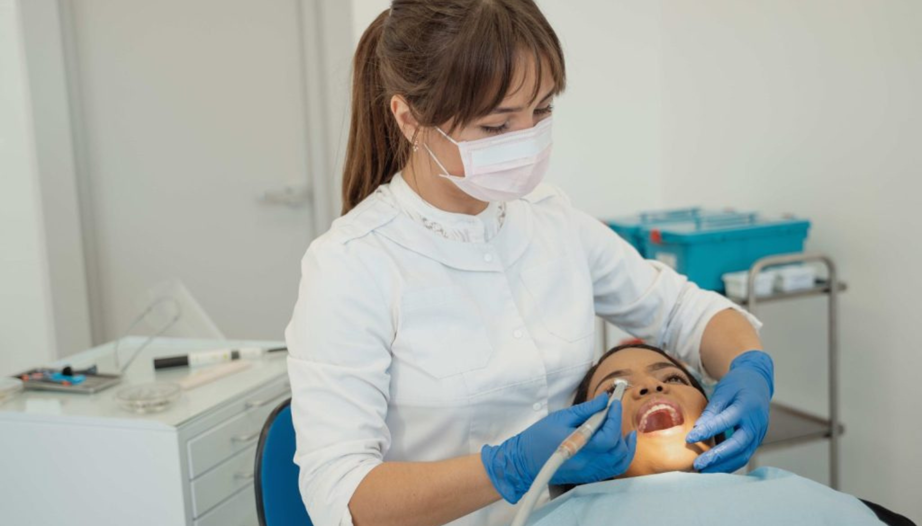 Tannlege med munnbind og blå hansker utfører tannrens på kvinnelig pasient som sitter i tannlegestol i en lys og moderne tannklinikk. 