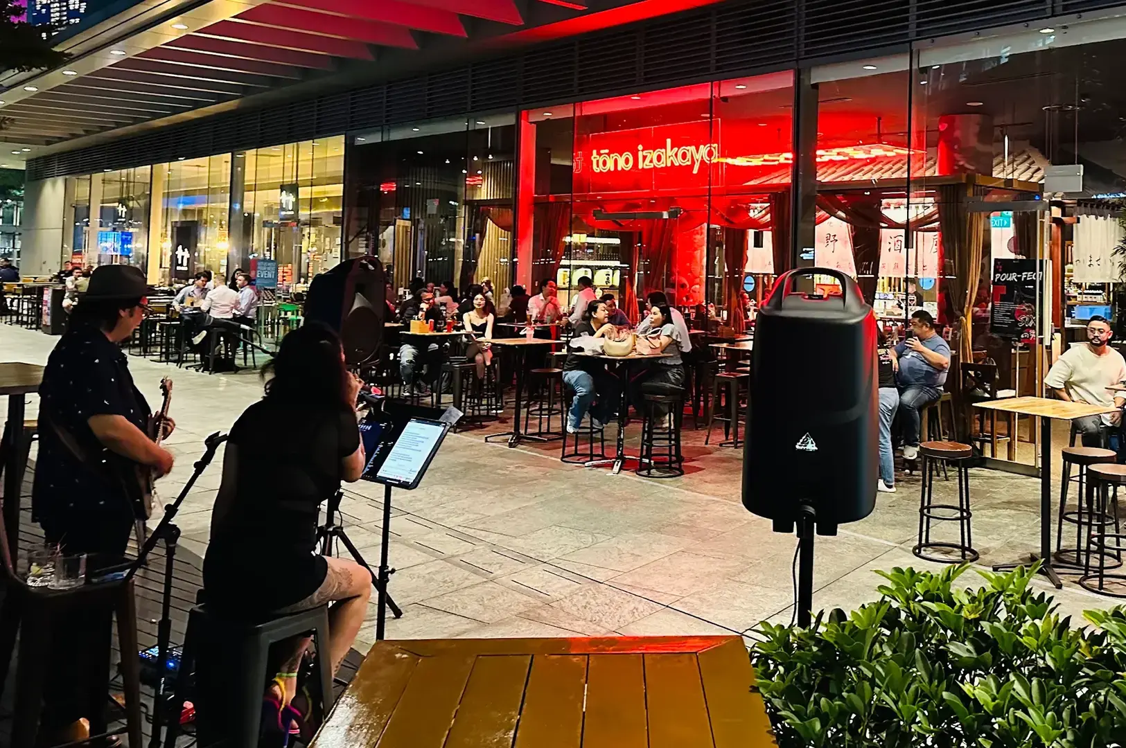 Tono Izakaya’s red-lit restaurant frontage with outdoor tables, seated diners, musicians performing in the foreground, and speakers set up along the mall walkway.