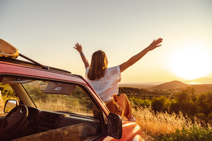 Happy young woman sitting on the hood of a car at sunset.