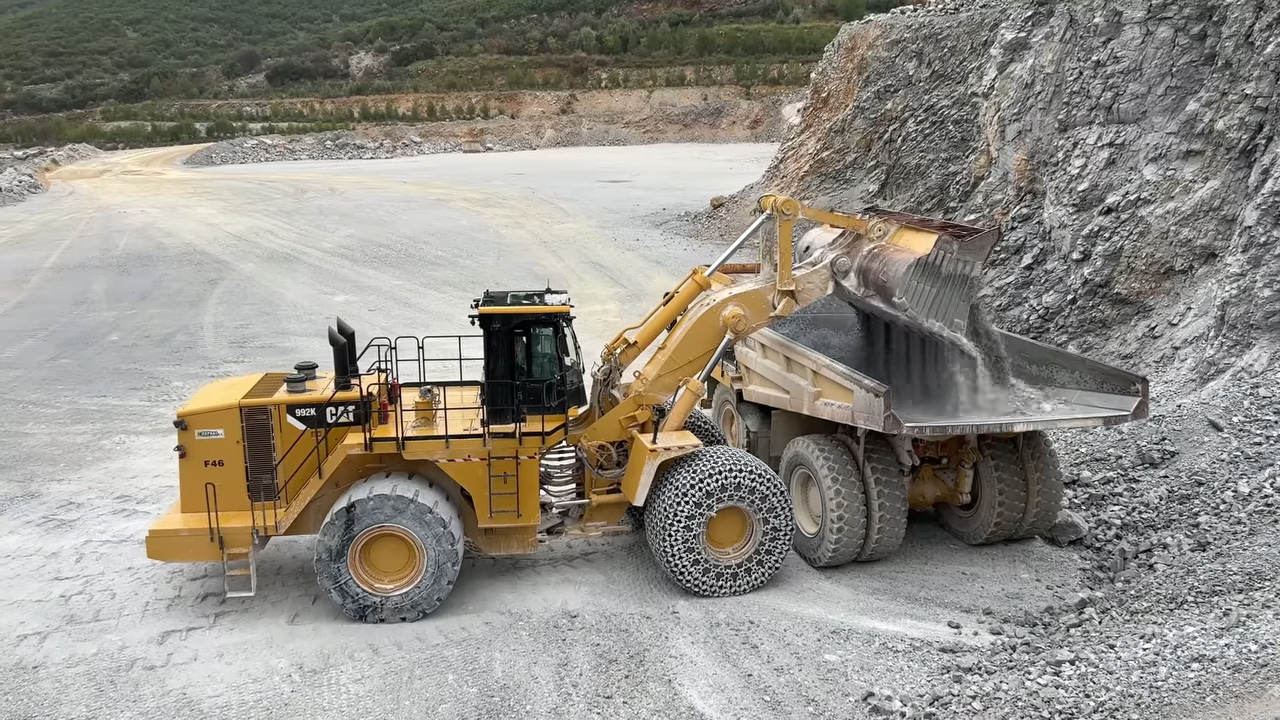 A yellow front-end loader transferring gravel into the back of a large dump truck at a quarry site with a rocky hillside in the background