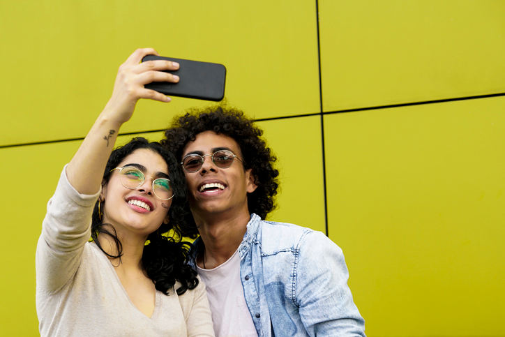 Happy young couple smiling for a selfie in front of a yellow wall. 