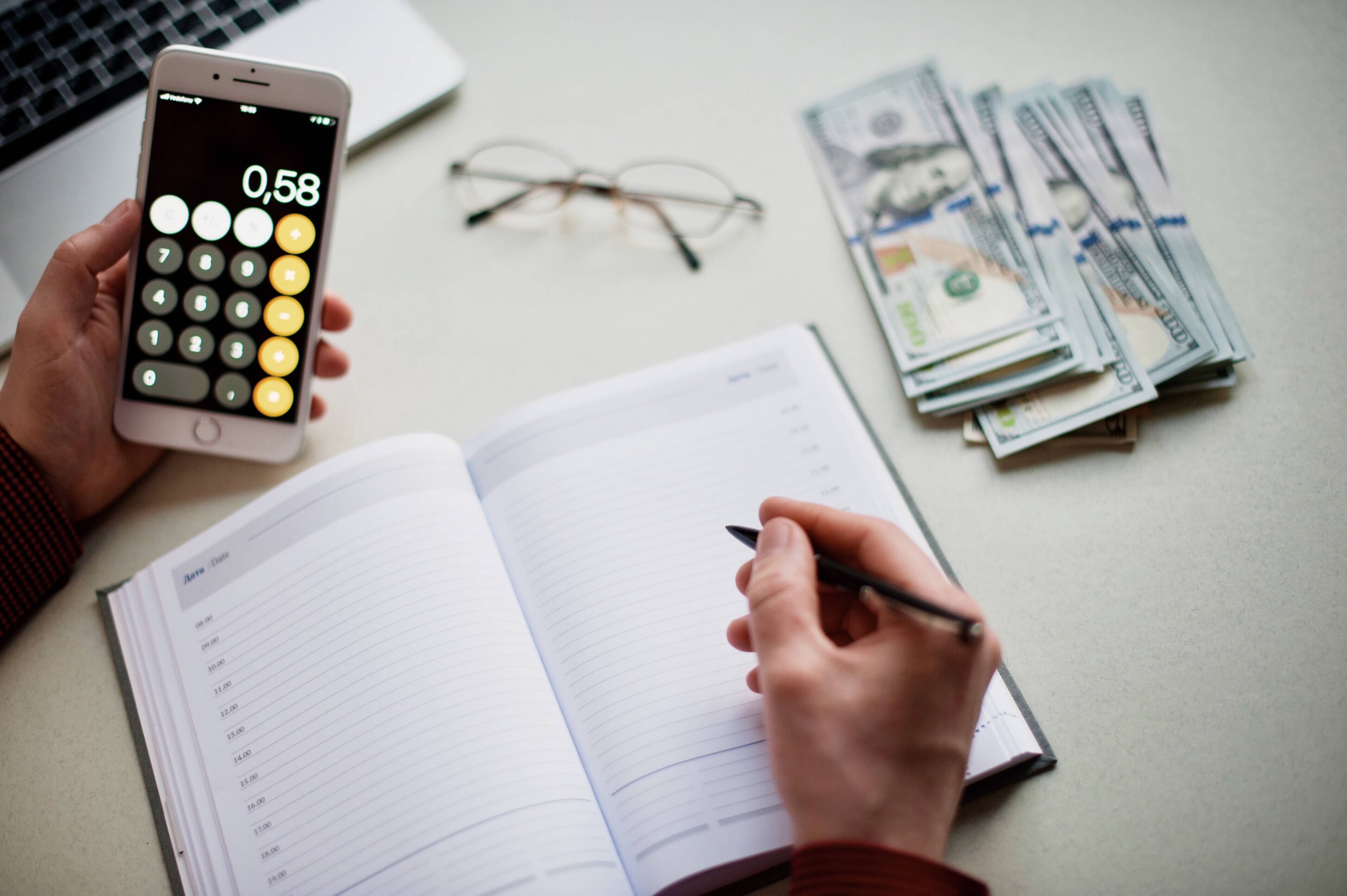 A person uses a smartphone calculator and writes in a planner next to a stack of cash, illustrating the process of organizing finances into essential budget categories.