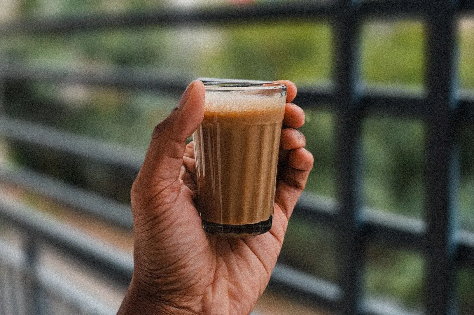 A close-up shot of a hand holding a small, clear glass filled with freshly poured, frothy masala chai, set against a softly blurred background of a grey metallic railing and greenery.