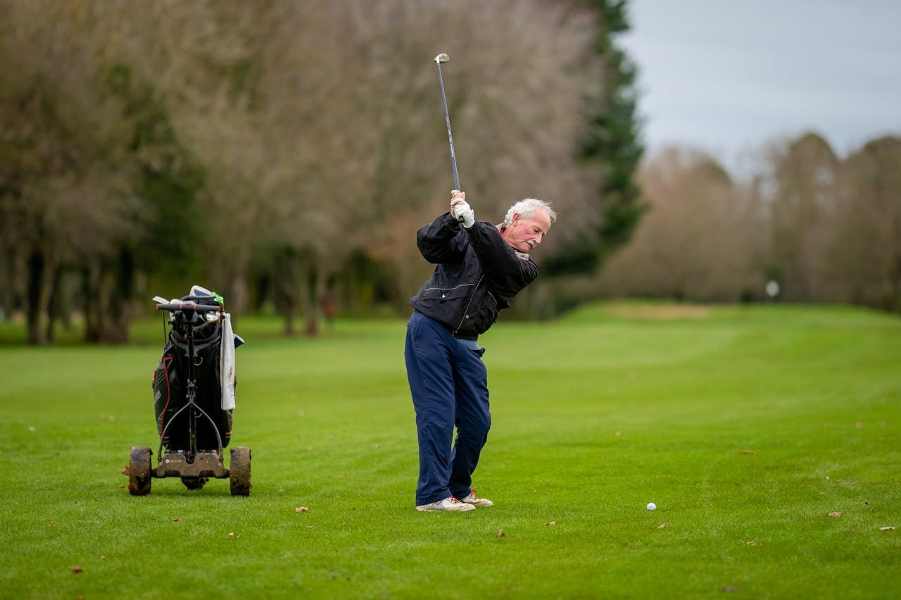 Senior golf fitness on the fairway, older golfer making a full swing near a push cart on a green course.