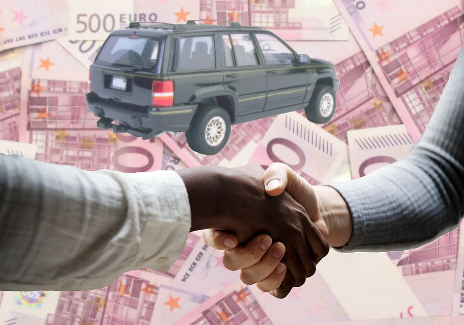 A man shaking hands with a car buyer, with a junk car in the backgrou
