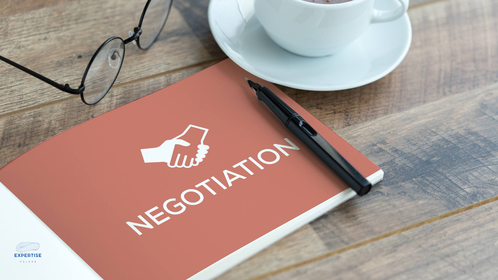negocjacje Otwarty notatnik z napisem 'NEGOTIATION' oraz grafiką dwóch dłoni uściskujących się, leżący na drewnianym stole. Obok znajduje się pióro, okulary oraz filiżanka kawy