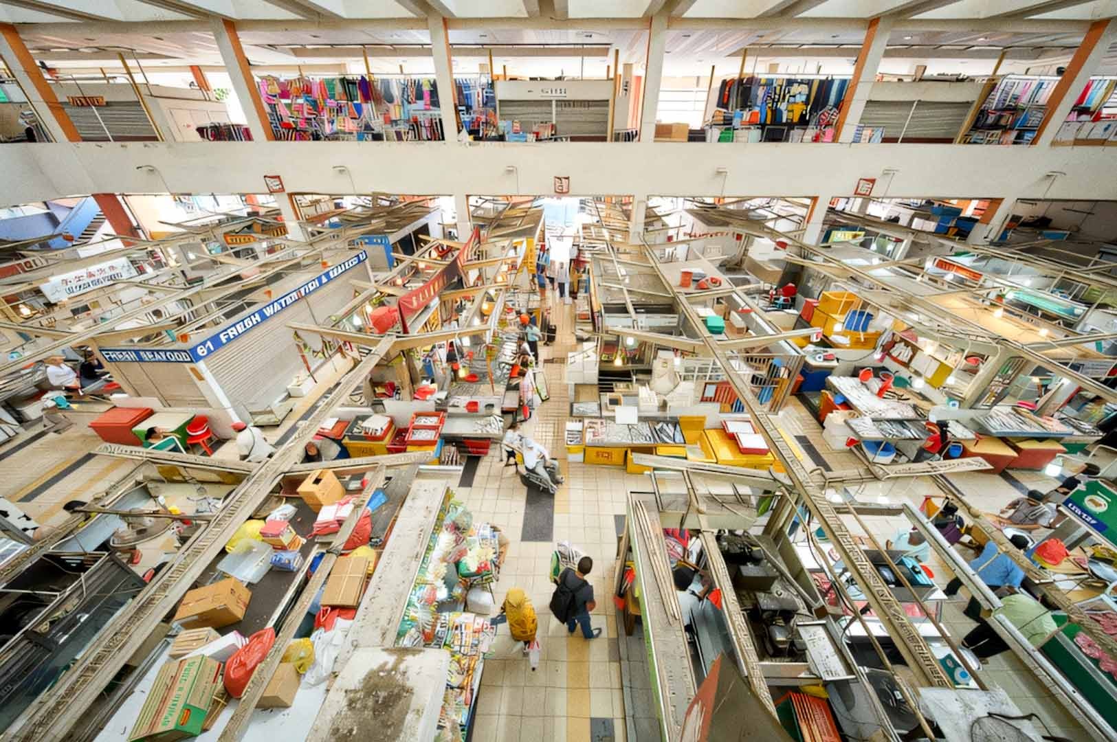 Indoor Tekka market in Singapore with vendor stalls selling fresh seafood, produce, packaged goods, and textiles, multi-level layout with shoppers walking through aisles.