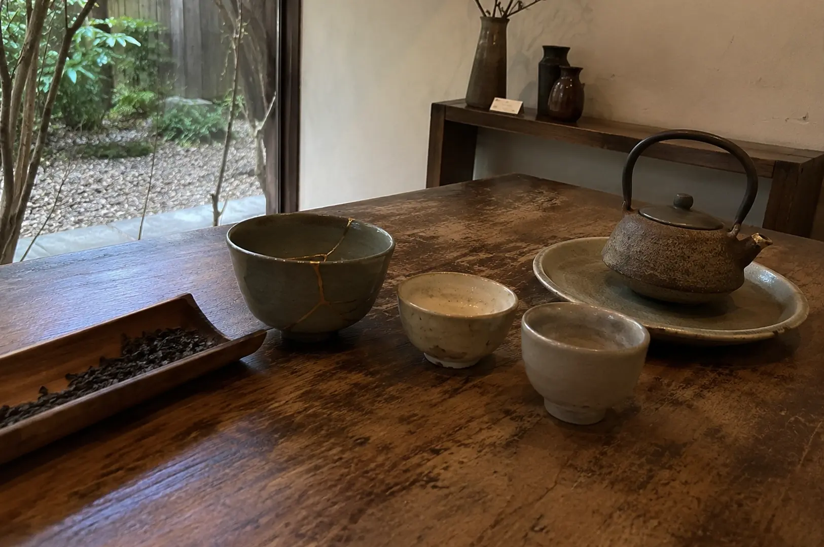 An elegant and minimalist tea arrangement is showcased on a weathered, textured wooden tabletop, highlighted by a large ceramic tea bowl beautifully repaired using the traditional Japanese art of kintsugi, with prominent gold lacquer mending its cracks. Beside the kintsugi bowl are two smaller, rustic ceramic teacups, a long curved wooden bamboo scoop holding dark, loose-leaf tea, and a dark, textured cast-iron teapot resting on a matching wide ceramic saucer. In the background, a large window reveals a tranquil stone and moss garden, while a simple wooden shelf displays minimalist ceramic vases against a plain wall.