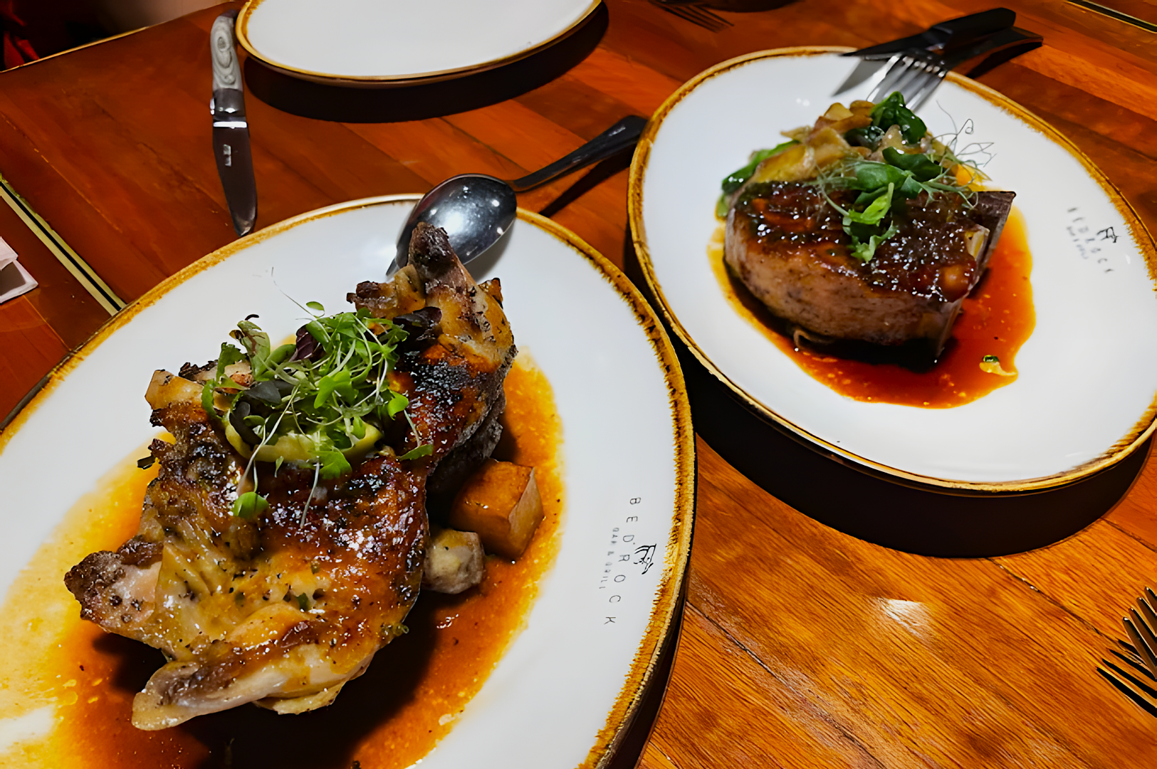 Grilled chicken with herbs, served with vegetables, on a white plate next to a beef steak in rich sauce with greens. Rustic wooden table setting.