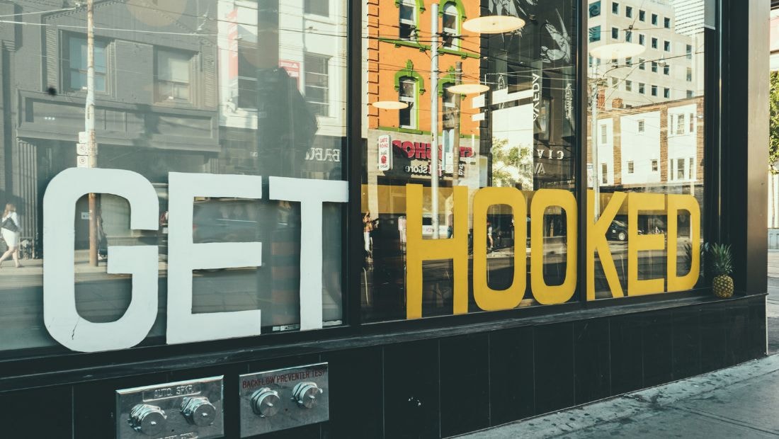 Large, bold storefront window display featuring the phrase โGET HOOKEDโ in oversized white and yellow letters, with reflections of city buildings and street activity visible in the glass.