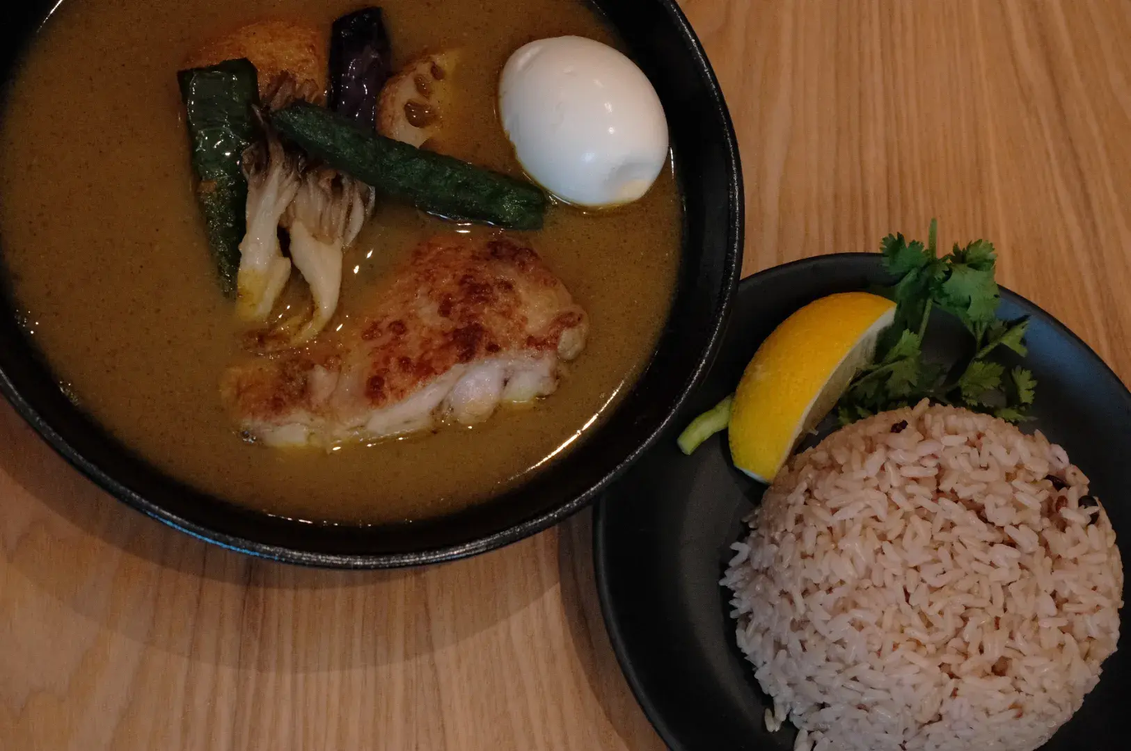 A bowl of curry with chicken, vegetables, and a soft-boiled egg, accompanied by a plate of brown rice and a lemon wedge.