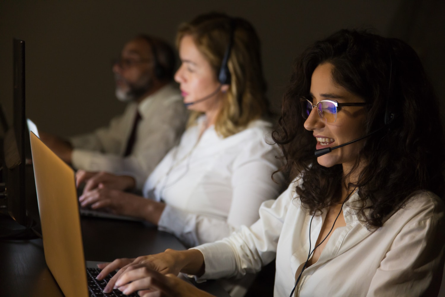 A dimly lit office shows three professionals working on their laptops, each wearing headsets. The focused atmosphere illustrates the importance of scalable contact center platforms in providing 24/7 omnichannel customer support.