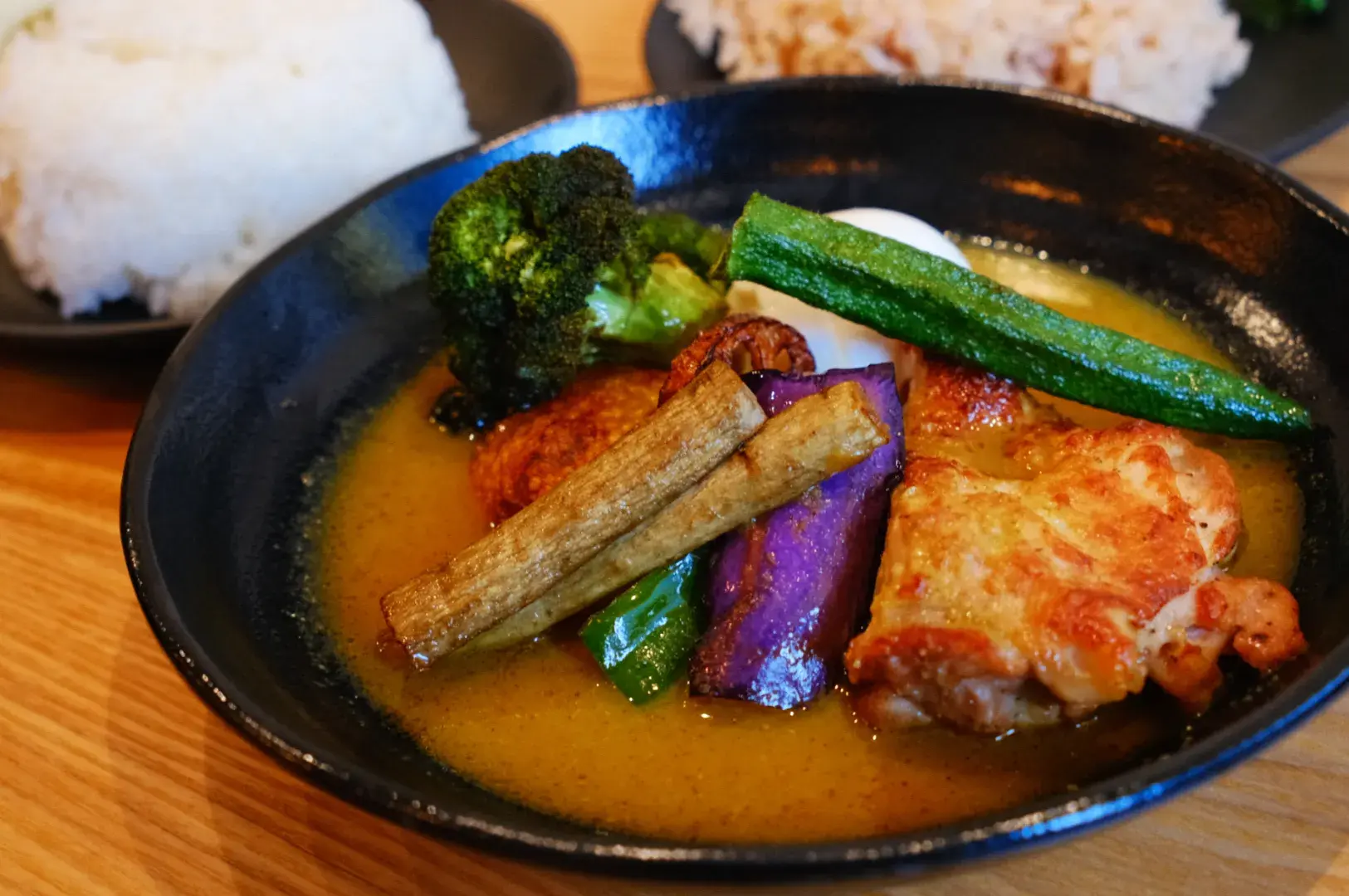 A bowl of curry featuring chicken, broccoli, okra, and eggplant, served with a side of rice.