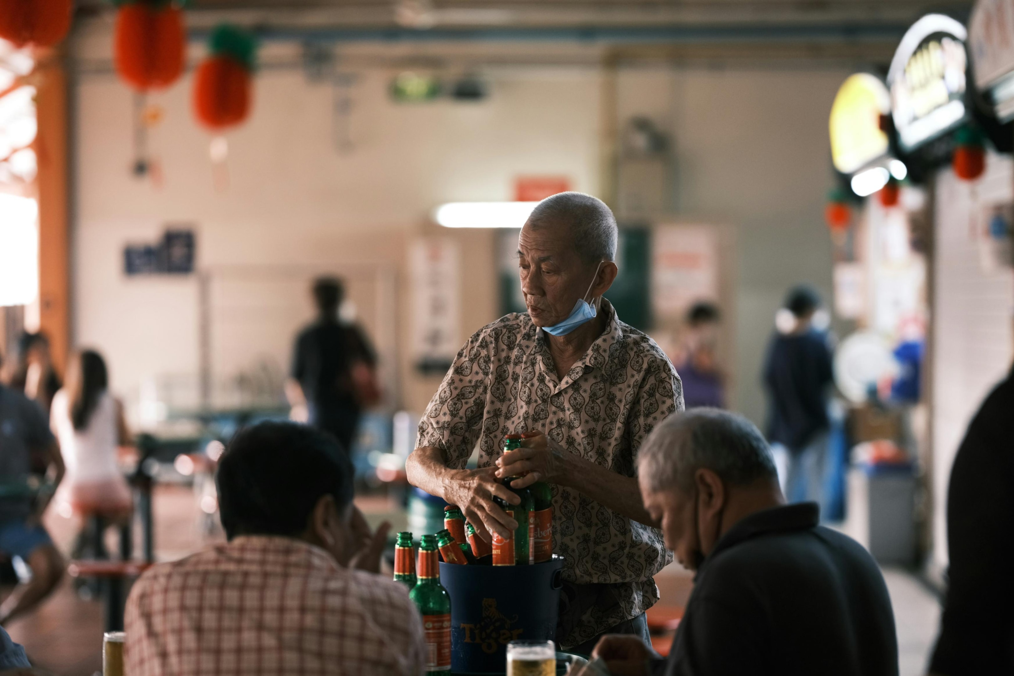  In a bustling open-air market, an elderly man wearing a face mask tucked under his chin carefully pulls green Tiger Beer bottles from a blue ice bucket to serve customers seated at a nearby table. The background remains softly blurred, showing other patrons and vibrant red lanterns that add a sense of local atmosphere to the scene.