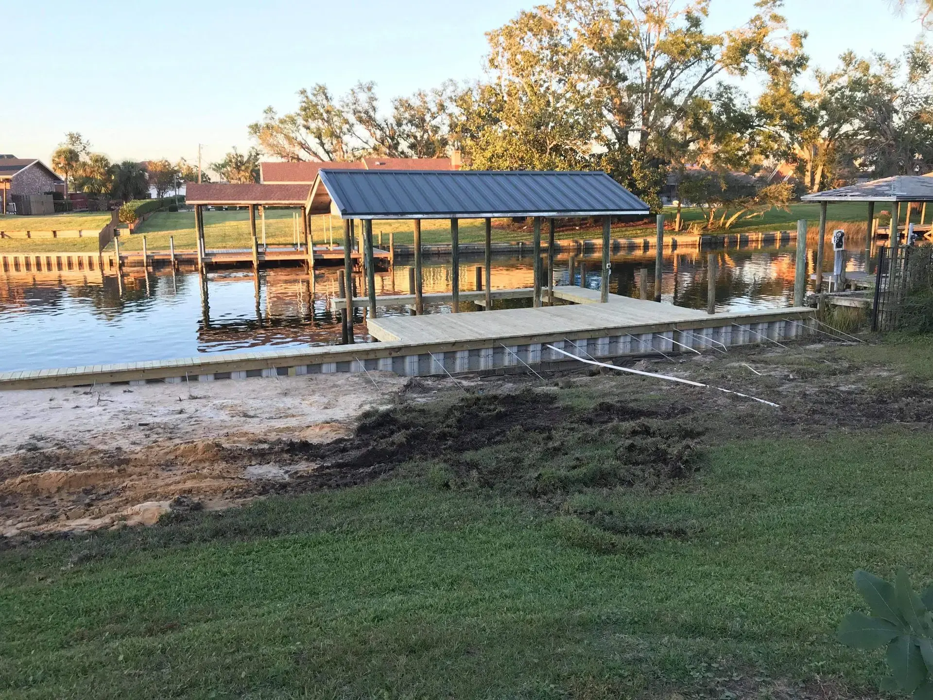 A newly constructed retaining wall and dock along a calm canal with grassy banks and houses in the background during sunset.
