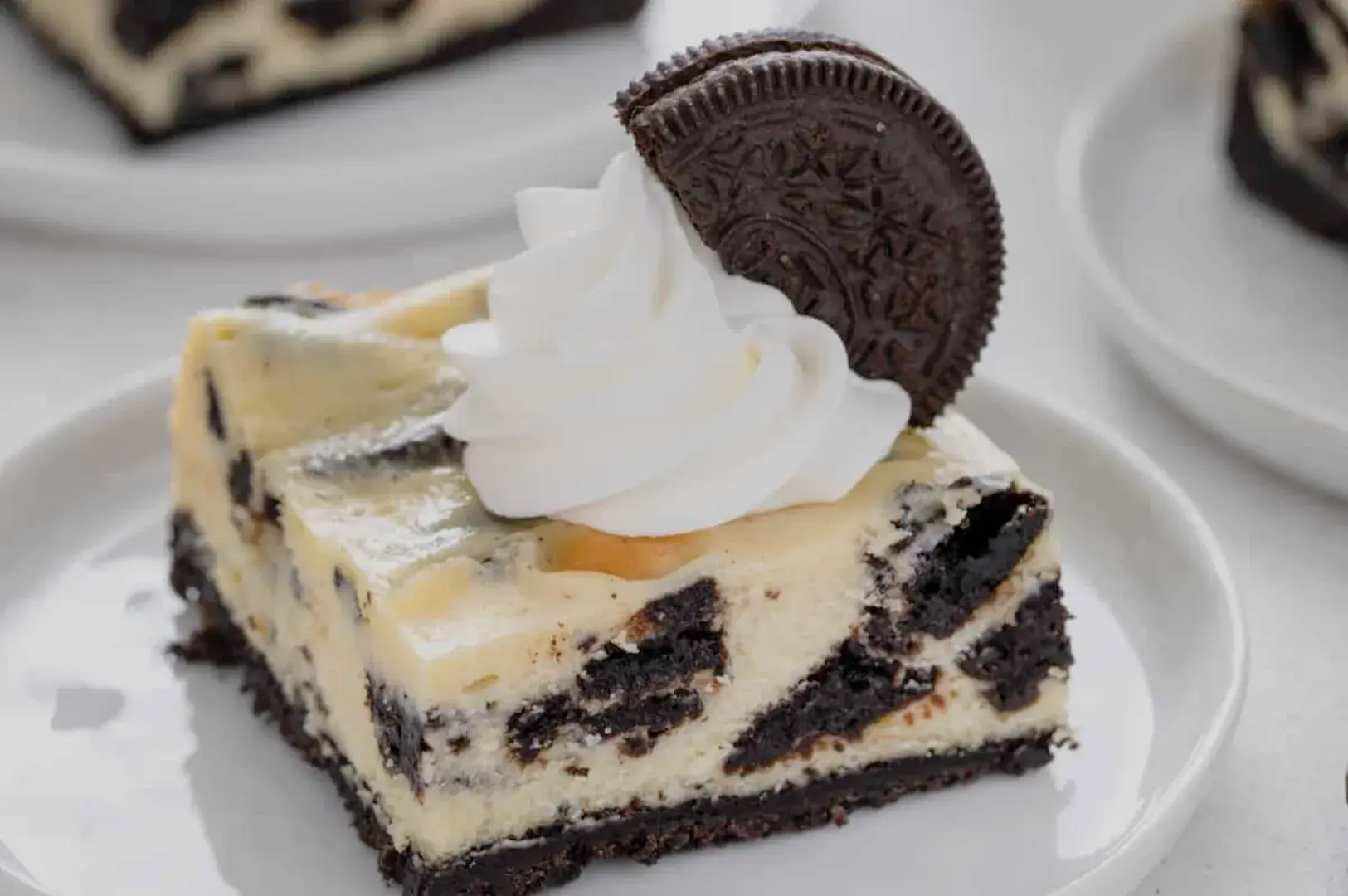A slice of cream-filled dessert topped with a dollop of whipped cream and an Oreo cookie, served on a white plate.