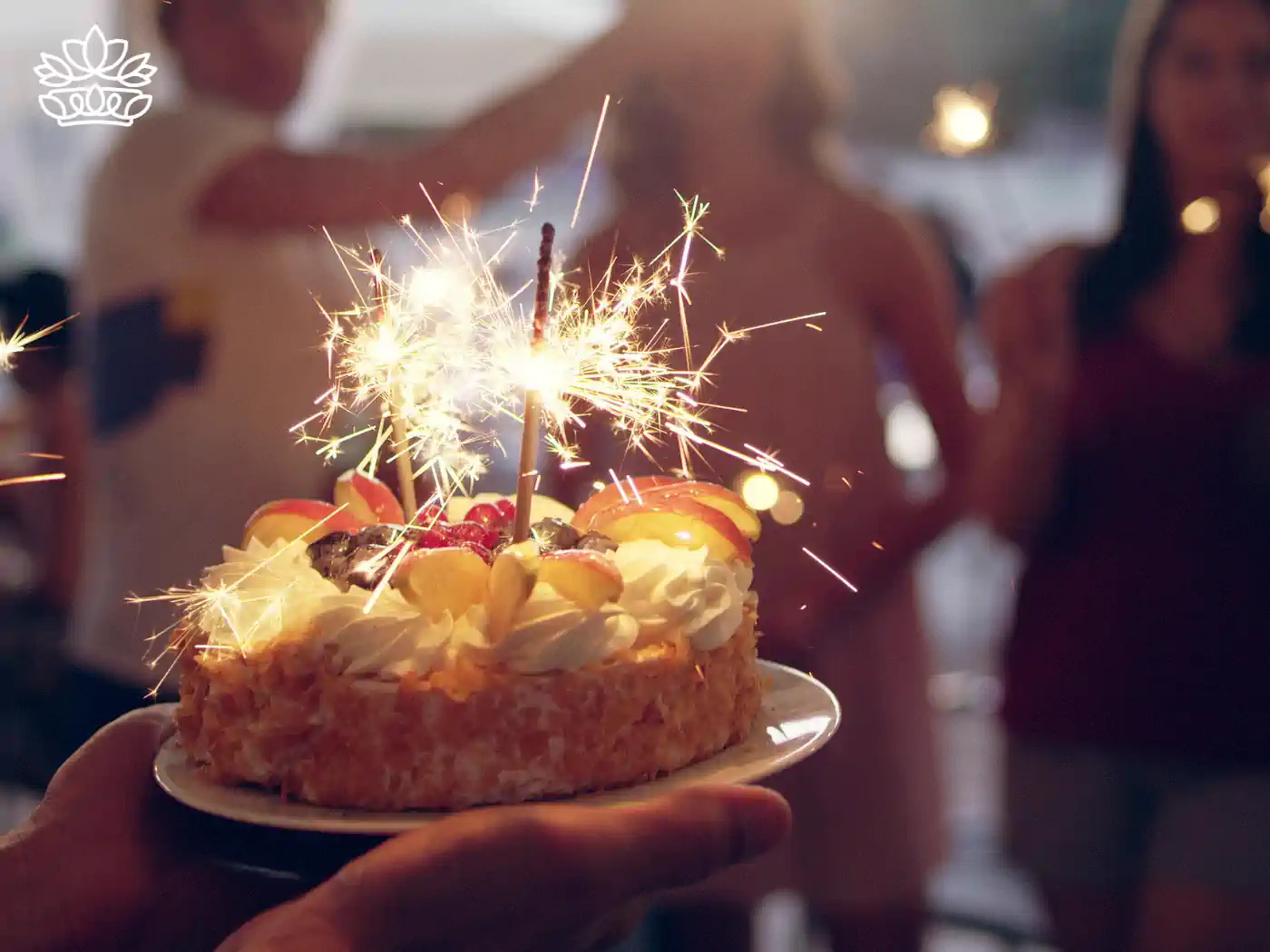 A birthday cake with sparklers being held at a party with people in the background. Fabulous Flowers and Gifts. Taurus Flowers & Gifts Collection.