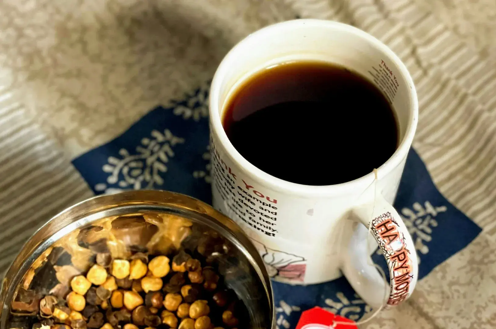 A high-angle shot featuring a white ceramic mug filled with dark tea, resting on a navy blue patterned fabric napkin next to a small stainless steel bowl filled with roasted chickpeas, set against a light, neutral-colored background.