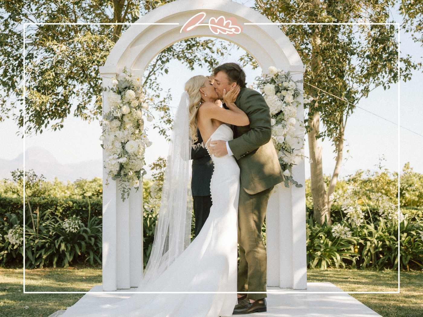 Tess and Greg sharing their first kiss as husband and wife under a white floral arch at their Nantes Estate wedding in Paarl