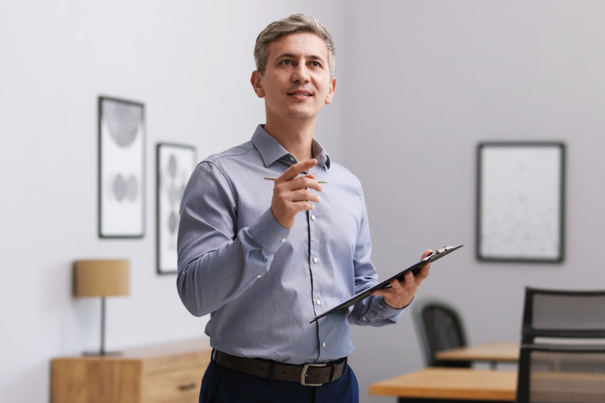 A man in a blue shirt holds a clipboard and a pen, looking up and to the side as if in thought.
