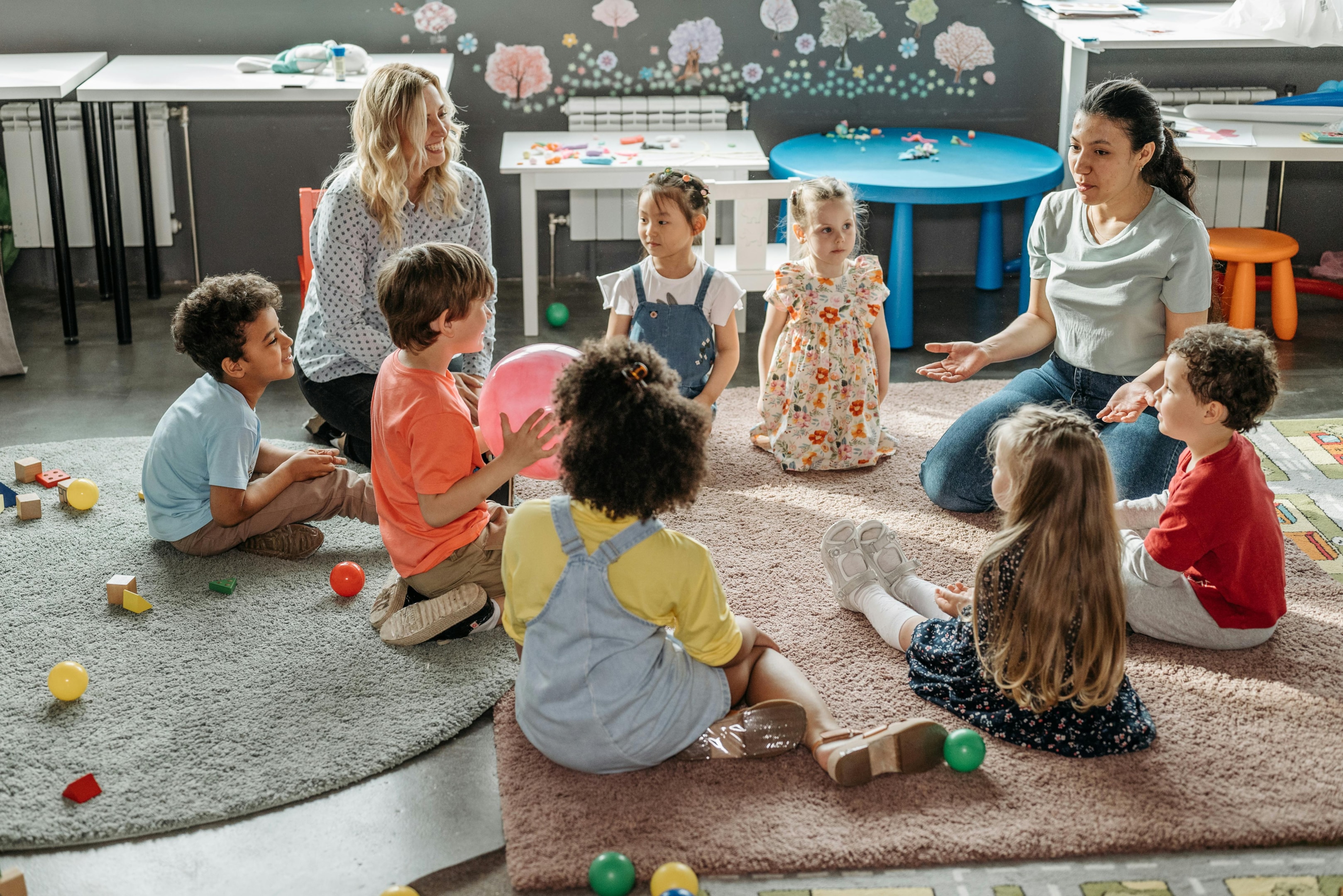 Nursery school children sitting in a circle with 2 teachers listening to one of them give instructions