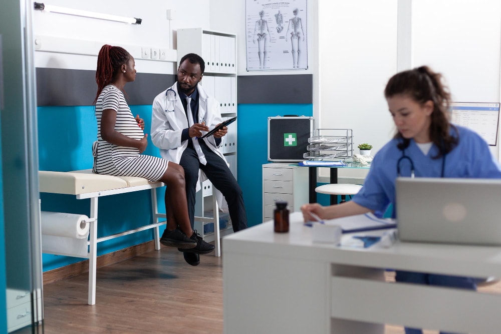 A male doctor speaks with a pregnant patient seated on an exam table while a nurse in the background uses a laptop at a desk. Unified Communications platforms and team collaboration tools help maintain seamless communication across roles, enhancing business productivity in outpatient care environments.