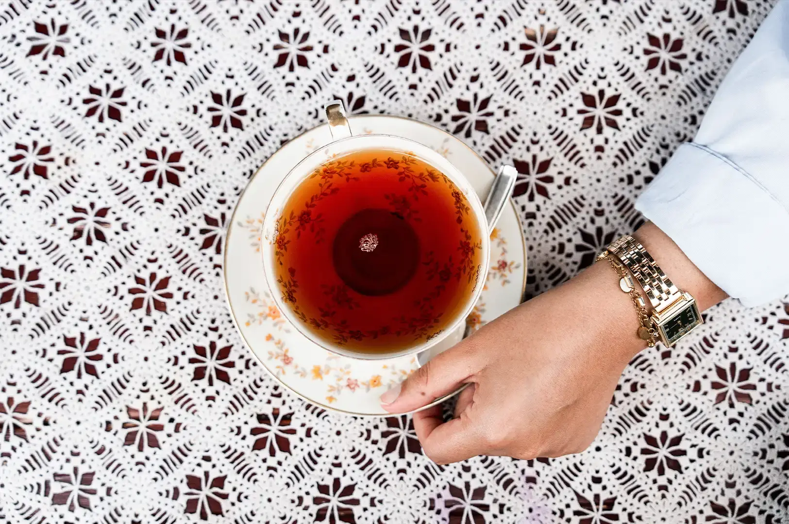 A top-down shot captures a person wearing a gold wristwatch holding the rim of a delicate floral porcelain teacup filled with dark tea, set against an intricate white lace tablecloth.