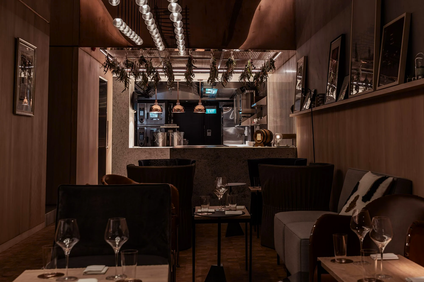 A dimly lit, cozy restaurant interior with elegant tables and dark armchairs. Decorative lights and hanging plants enhance the warm, intimate ambiance.