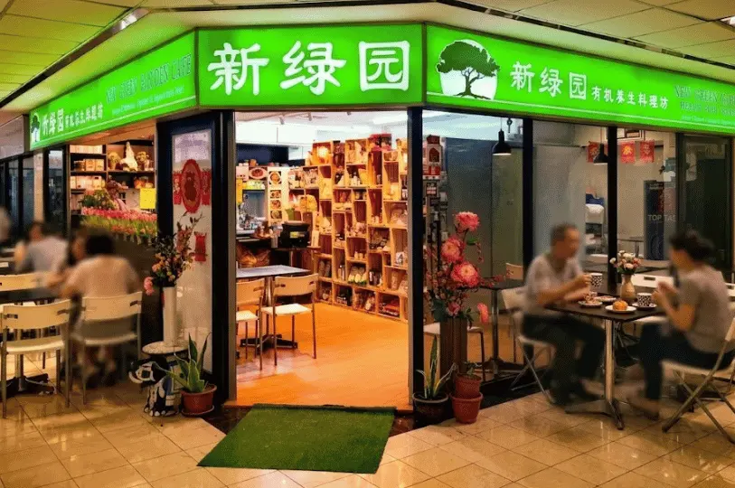 The brightly lit green storefront of Green Pasture Cafe, a vegetarian eatery, with patrons dining at tables outside the shop's entrance.