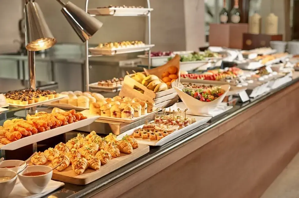 A long buffet table is neatly arranged with an extensive variety of breakfast items, including fresh croissants, pastries, and savory appetizers under warm heat lamps. The spread also features wooden crates of whole fruit, bowls of vibrant berry salad, and tiered trays of desserts in a bright, modern dining setting.