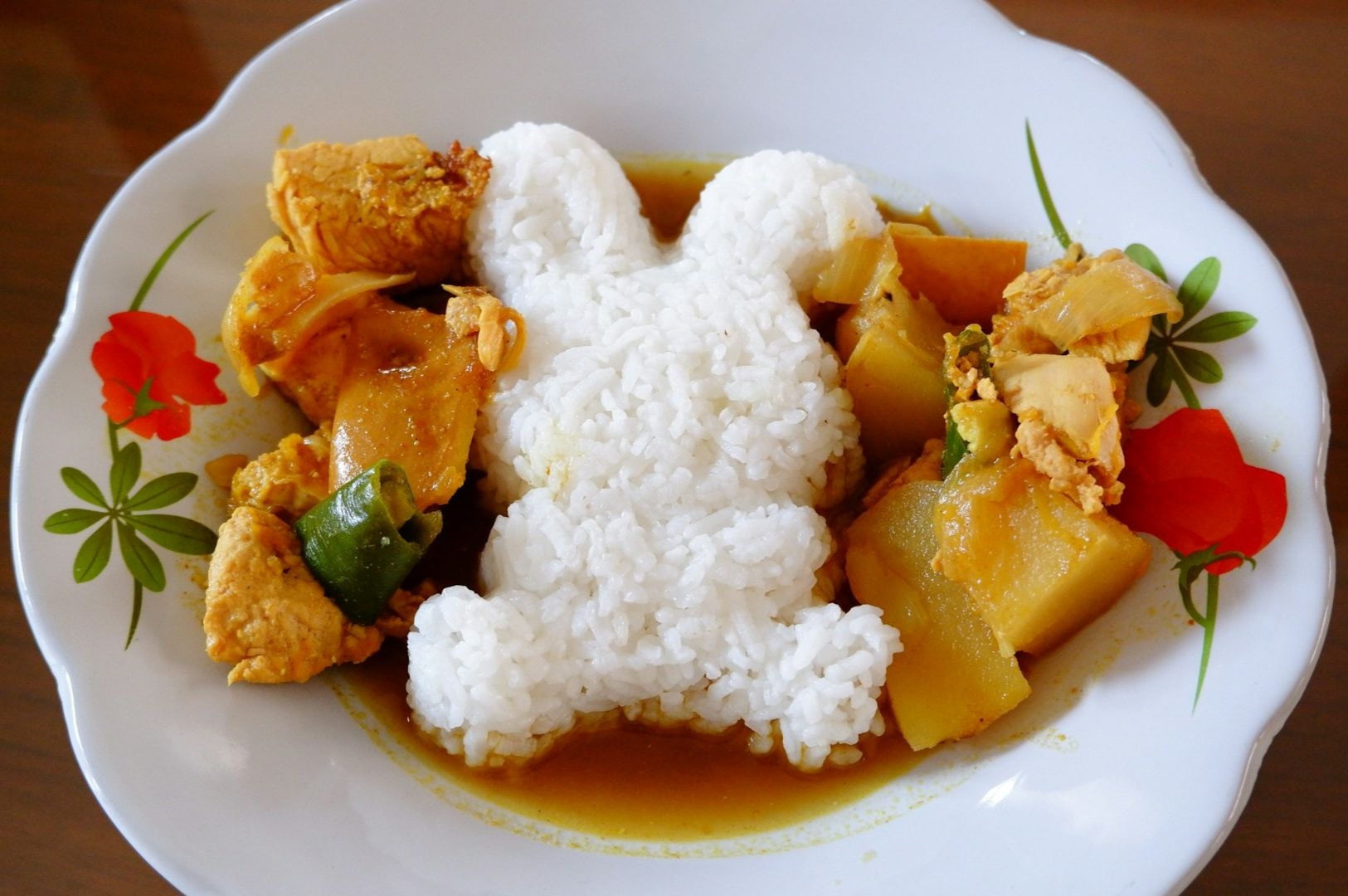 A plate of curry with chicken and potatoes surrounds rice shaped like a bunny. The dish is garnished with red flower designs, creating a playful, appetizing look.