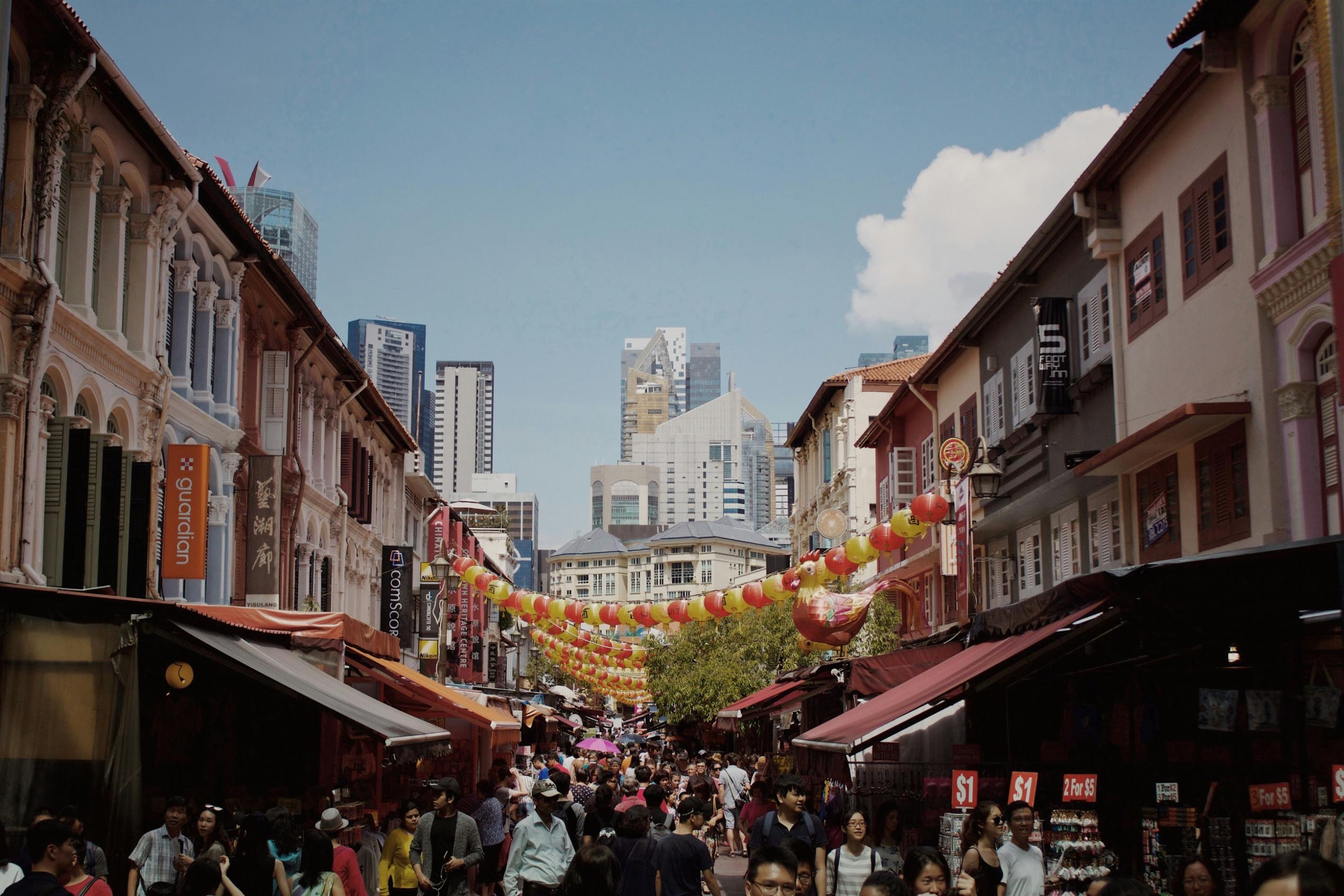 This image presents a vibrant street view, likely in Singapore or a similar urban hub, where crowds of people are walking below strings of red and yellow lanterns that span across a narrow street lined with traditional shop houses and market stalls.