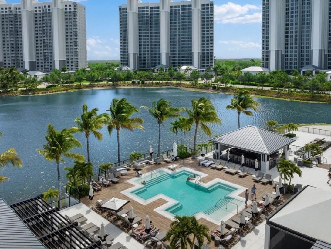 The image showcases the expansive clubhouse amenities of the prestigious Kalea Bay community, featuring tennis courts, resort-style pools, and a wellness center, all surrounded by lush tropical landscaping. This vibrant setting embodies the essence of luxury coastal living in Naples, FL, offering residents a sophisticated retreat with world-class amenities.