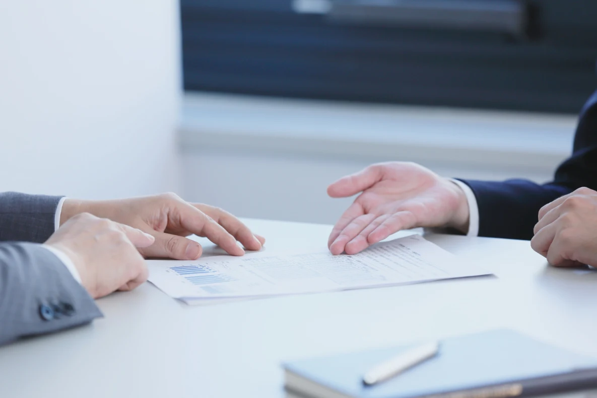 Two individuals in business attire review a document with charts on a white table, accompanied by a notebook and pen.