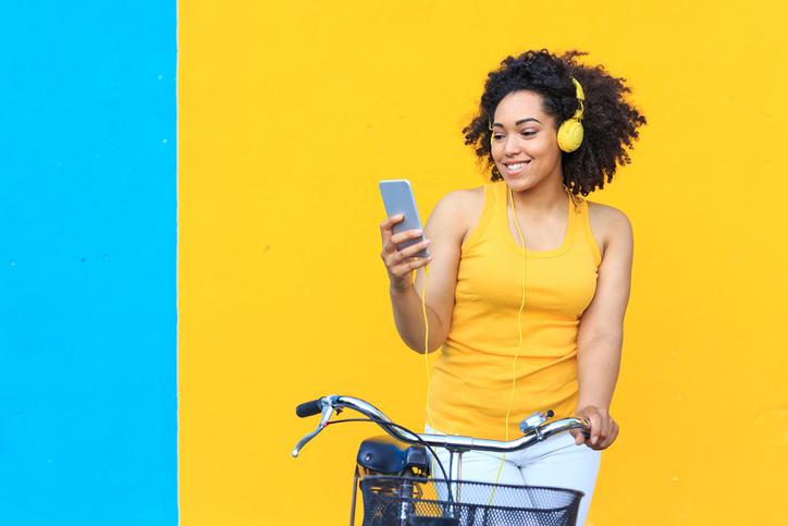 Happy young woman in a yellow tank top with yellow headphones listening to music on her cell.