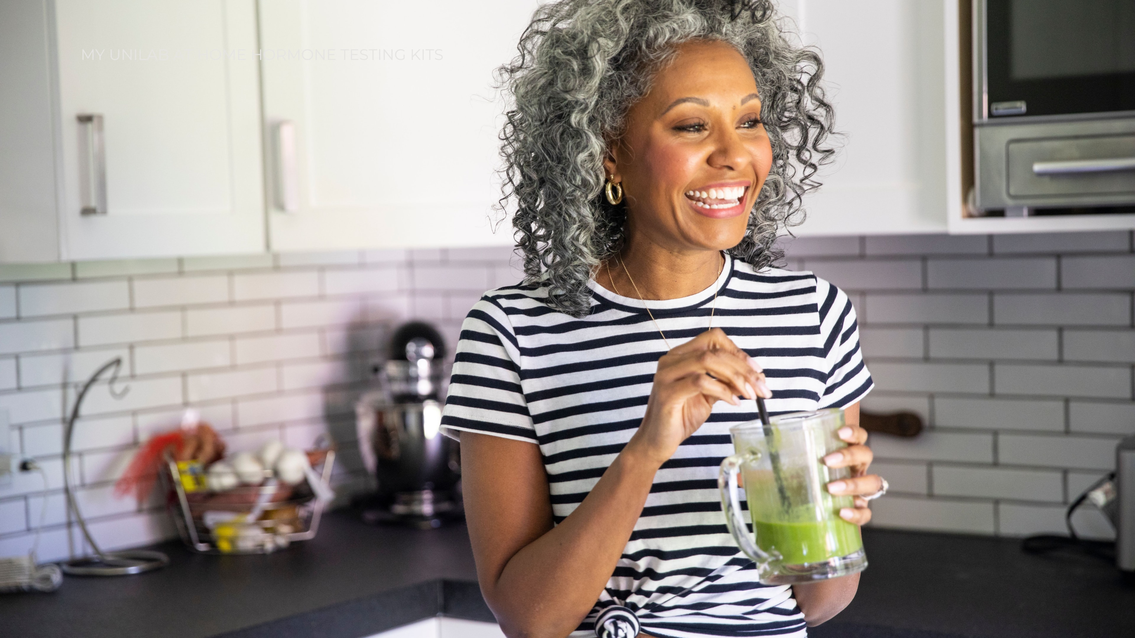 A middle aged female in her kitchen smiling while drinking a fresh green juice.